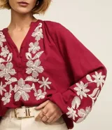 Blusa color borgoña de batista con cuello en V y botones. Presenta bordados florales blancos en el frente y las mangas largas con puños elásticos.