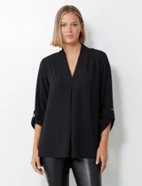 Blusa negra de manga larga con escote en V y pliegue central. La tela presenta un tejido piqué texturado sutil. Las mangas se pueden remangar y sujetar con una presilla y botón dorado.