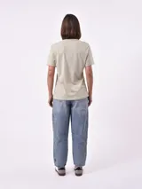 Remera blanca de algodón con cuello redondo y manga corta. Presenta un pequeño bordado en el pecho izquierdo con una figura que parece ser una persona acostada en una tabla de surf o flotador.