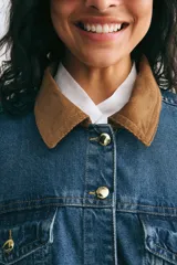 Campera de denim azul con cuello de pana marrón, botones metálicos dorados, canesú delantero y trasero, corte holgado, hombros caídos, puños abotonados, bolsillos laterales discretos y bolsillos superiores con solapa y botón.