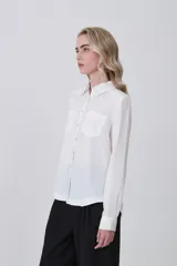 Camisa blanca de manga larga con cuello clásico, dos bolsillos frontales con tapa y botones a presión. Presenta un tejido con textura sutil de rayas verticales.