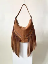 Bolso tipo hobo color tostado, hecho de cuero con flecos largos en el frente y dos bolsillos con cierre. Tiene una manija regulable con hebillas metálicas.