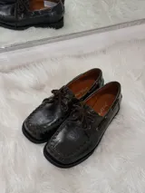 Zapato tipo mocasín de cuero color chocolate, con suela plana de goma negra, cordones al tono y costuras decorativas visibles en el borde superior.