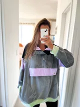 Buzo oversize gris con bloques de color negro, lila y verde. Tiene cuello alto con cierre de botones a presión y bolsillo tipo canguro con solapa abotonada.