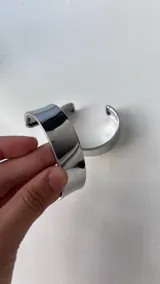 Pulsera tipo brazalete o cuff de metal plateado, con acabado liso y brillante, de diseño abierto.