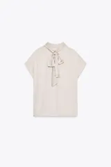 Blusa color crudo de tejido satinado, con cuello subido, manga sisa y cierre en la espalda con lazada y botones.