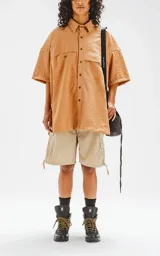 Camisa manga corta color beige, con cuello clásico, cierre frontal con botones, doble bolsillo con ojal en el delantero y silueta oversize.