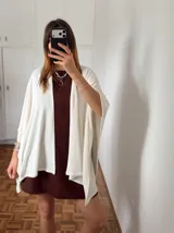 Ruana o poncho de tejido fino color crema o blanco roto, de corte amplio y asimétrico, sin cierres ni cuello definido.