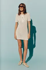 Vestido corto de tejido pointelle color crema, con cuello redondo, mangas cortas y abertura frontal con dos moños decorativos.