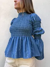 Camisa celeste con estampado de flores blancas, cuello alto con volantes, mangas largas abullonadas con puños elásticos y peplum.