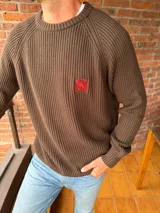 Sweater de tejido de punto color beige, con cuello redondo y manga raglán. Presenta un pequeño bordado en hilo rojo en el pecho.