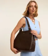 Cartera hobo grande de poliuretano color marrón oscuro con textura y correa de hombro. Tiene detalles de hebillas metálicas y bolsillos internos separados.