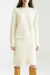 Conjunto tejido color crema, compuesto por un sweater de mangas largas con diseño trenzado y falda midi con diseño trenzado y flecos en el ruedo.