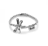 Pulsera elástica plateada con cuentas redondas y dije de libélula. Incluye un dije rectangular con la palabra "Suerte" grabada y un dije cilíndrico con el logo "UNO de 50".