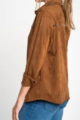 Camisa marrón de gamuza vegana con cuello clásico, cierre frontal con botones y mangas largas con puños abotonados. Presenta detalles de tachas metálicas en cuello y charreteras.