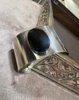Brazalete rígido de alpaca plateada con piedra ovalada negra engarzada en el centro.