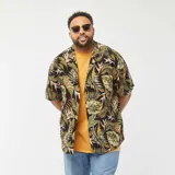 Camisa de manga corta con estampado de hojas tropicales en tonos verdes y marrones sobre fondo negro. Tiene cuello clásico, corte recto y cierre frontal con botones.