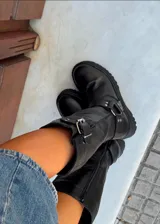 Bota de caña alta color negro, realizada en cuero encerado, con hebillas en la caña y herrajes de níquel. El fondo está realizado artesanalmente, es de suela, y es negro. El interior está forrado en forrería vacuna.