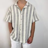 Camisa de manga corta, cuello cubano, color crudo con rayas verticales finas en gris oscuro. Presenta un bolsillo de parche en el pecho y botones marrones.
