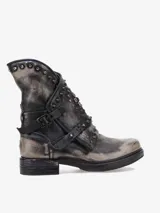 Bota corta de cuero bovino, estilo militar o rockero, con acabado desgastado en tonos grises y negros. Presenta detalles de tachas metálicas en el borde superior y correas ajustables con hebillas y remaches decorativos.