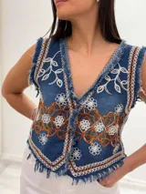 Chaleco corto de denim azul con bordados florales en blanco y marrón, detalle desflecado en escote, sisas y ruedo. Escote en V y cierre frontal con botones. Estilo boho chic.