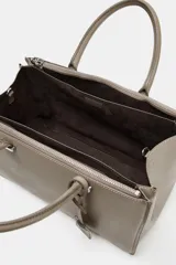Bolso de mano tipo satchel o tote pequeño, color topo o gris pardo, con doble asa superior y cierre principal por cremallera. Presenta un colgante rectangular de cuero sintético colgando de una de las asas.