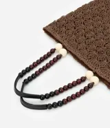 Bolso de mano tipo tote, tejido en punto de ganchillo con textura de conchas, color marrón oscuro. Las asas son de cuero o material sintético oscuro, adornadas con cuentas redondas de madera en tonos marrón oscuro, borgoña y crema.