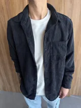 Sobrecamisa de pana negra con cuello clásico, cierre frontal con botones, manga larga con puños abotonados y bolsillo de parche en el pecho.