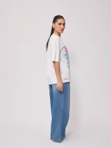 Remera blanca de algodón con estampado frontal de estilo playero con texto "Welcome to the Surf Camp" y dibujo de una casa de playa con palmeras y tablas de surf.