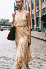 Vestido midi de cuello halter con estampado paisley en tonos beige, amarillo y marrón. Tiene lazada en la espalda, cintura elástica y falda plisada. Cierre trasero con cremallera oculta, abertura y botón.