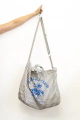 Bolso de tela tipo tote bag, con estampado de rayas verticales en tonos beige y negro/gris. Presenta un estampado frontal en azul que incluye una figura ecuestre bajo una palmera.