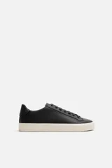 Zapatilla urbana de cuero sintético color negro, con cordones y suela de goma color crema.