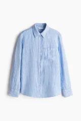 Camisa celeste y blanca a rayas verticales, de lino liviano, con cuello clásico, botones frontales, canesú de doble capa en la espalda, hombros caídos, mangas largas con puños abotonados, bolsillo de parche en el pecho y bajo redondeado.