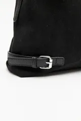 Cartera de gamuza color marrón, de textura suave y acabado aterciopelado. Cuenta con una silueta amplia y flexible, cierre superior metálico, correa regulable y detalles laterales con hebillas.