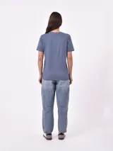 Remera blanca de algodón con cuello redondo y manga corta. Presenta un pequeño bordado en el pecho izquierdo con una figura que parece ser una persona acostada en una tabla de surf o flotador.