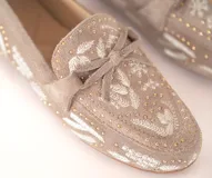 Mocasín de cuero gamuzado color avena, con bordados contrastantes, apliques metálicos y lazo decorativo en la capellada.