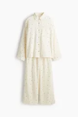 Pijama de dos piezas en muselina de algodón color crema con estampado de pequeñas figuras oscuras. Incluye una camisa de manga larga con cuello camisero, botones frontales y bolsillo de parche, y un pantalón recto con cintura elástica y cordón de ajuste.