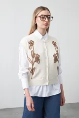Chaleco de punto color blanco con bordado aplicado de cordón marrón en diseño floral. Presenta cierre frontal de botones al tono, cuello redondo y terminaciones acanaladas en sisas y ruedo.