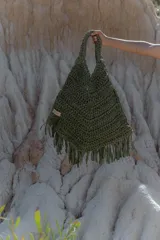 Bolso de mano grande tejido a crochet con técnica de trapillo reciclado, color verde oliva. Presenta un diseño amplio con asas trenzadas y flecos largos en el borde inferior. Tiene una pequeña etiqueta de cuero con el logo de la marca.