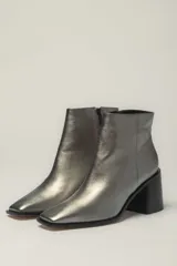 Botín de cuero metalizado color peltre con punta fina y cierre lateral. Tiene taco geométrico de madera color negro.