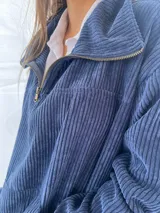 Buzo de pana fina color verde oliva, con diseño de corte holgado y mangas largas. Presenta una textura de cordón vertical característica de la pana y un pequeño parche decorativo en la parte superior de la espalda.