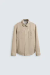 Sobrecamisa color beige de tejido con elasticidad, cuello solapa, manga larga con puño abotonado, bolsillos de plastrón en el pecho y cierre frontal con botones a presión.