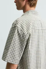 Camisa de hombre de corte boxy fit, confeccionada en tejido de algodón a cuadros en tonos beige, verde y azul. Tiene cuello solapa, manga corta y cierre frontal con botones.