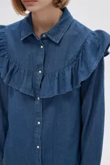 Camisa de jean azul con cuello clásico, cierre frontal con botones y mangas largas. Presenta volados en el canesú delantero y trasero.