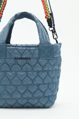 Cartera tote azul de nylon matelassé con diseño de corazones. Tiene manijas cortas y dos correas desmontables, una de cuero sintético y otra de tela trenzada multicolor. Cuenta con cierre superior y dos bolsillos internos.