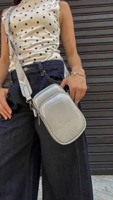 Bandolera pequeña tipo 'phone bag' de símil cuero color rosado pálido, con cierre de cremallera y correa ancha de tela con diseño de chevron en tonos claros.