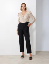 Enterito de fiesta con torso de lentejuelas color champagne, escote en V cruzado, mangas 3/4 con frunces en los hombros, cintura elastizada con lazo negro y pantalón negro de corte recto con bolsillos laterales.