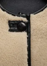 Chaqueta beige de poliéster con textura de borreguito, cuello redondo, cierre frontal con botones tipo trenca, ribetes en contraste de color negro, dos bolsillos delanteros y forro interior.