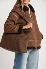 Chaqueta tipo gamulán de calce oversize en color marrón, con cuello y ribetes de corderito en contraste. Presenta cierre frontal de cremallera, bolsillos laterales y detalles de tiras decorativas en material sintético.