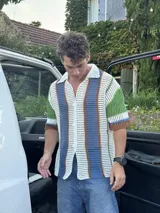 Camisa de manga corta tejida a crochet, con cuello polo y botones frontales. Presenta un diseño de rayas verticales en blanco, azul, verde y marrón.